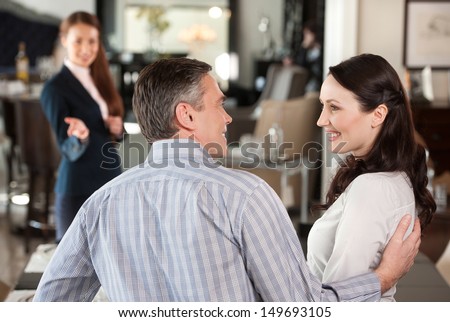 Mature Couple In Furniture Store. Cheerful Middle-Aged Couple Making Decision About Buying Furniture While Female Sales Clerk Waiting On Background