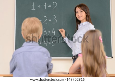 Young confident teacher. Confident young teacher standing near the blackboard and smiling while pupils sitting at desk on foreground