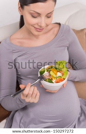 Pregnant woman eating salad. Smiling pregnant woman sitting on couch and eating salad