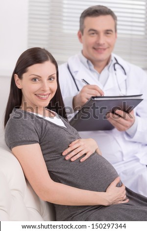 Medical exam. Confident mature doctor sitting near pregnant woman and making notes at his note pad