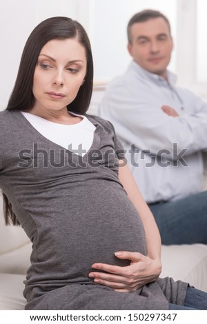Depressed pregnant woman. Depressed pregnant woman sitting on the couch and looking away while her husband sitting with his arms crossed on background
