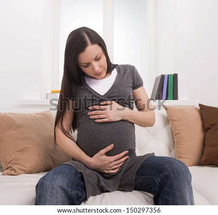 Waiting for a baby. Mature pregnant woman sitting on couch and holding hands on belly