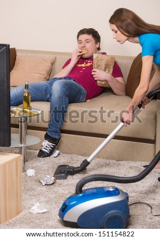 Mess at home. Young man eating potato chips while his girlfriend cleaning room