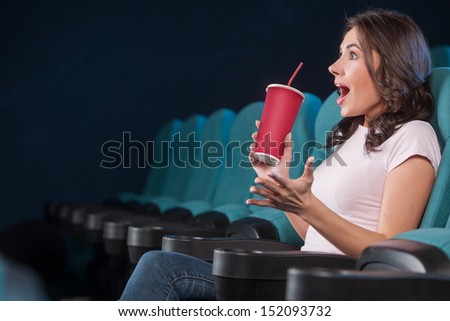Excited women at the cinema. Side view of beautiful young women drinking soda and gesturing while watching movie at the cinema