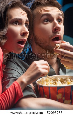 Exciting movie. Close-up of shocked young couple eating popcorn and drinking soda while watching movie at the cinema