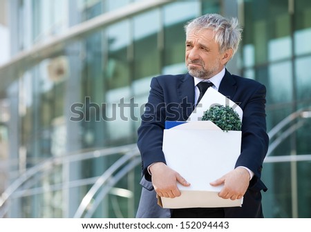 Fired. Depressed senior man in formalwear holding box with his work stuff and looking away