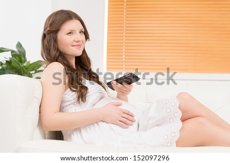 Pregnant woman watching TV. Beautiful pregnant woman watching TV while lying on the couch