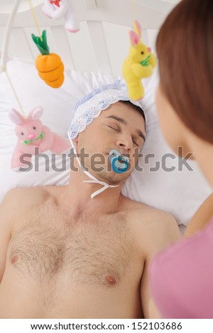 Big baby in bed. Top view of infant adult man lying on the baby bed while young woman looking at him