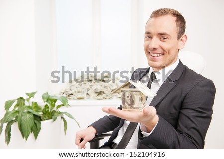 Wealthy man. Happy young man holding a house made from paper currency while a stack of money lying on the window sill