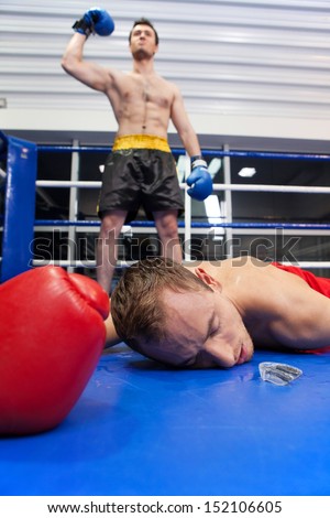 Winner and loser. Confident young boxer keeping his arm raised while his opponent lying down on the boxing ring
