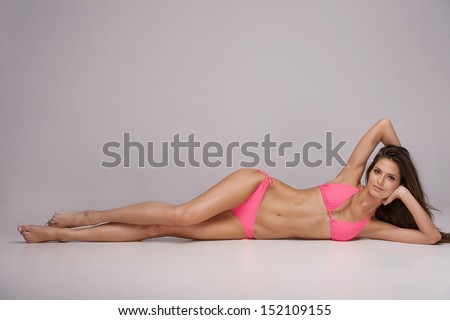 Beauty in pink bikini. Attractive young woman in bikini lying on side and looking at camera while isolated on grey