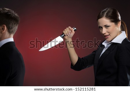 Wrong business partner. Smiling young woman in formalwear holding a knife near man in suit standing back to her