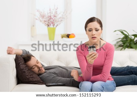 Checking his messages. Depressed young woman sitting near her sleeping boyfriend and looking at the mobile phone