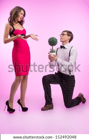 Nerd and beauty. Young nerd man standing at his knee and holding flower while beautiful young woman in red dress looking at him