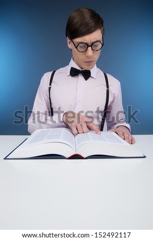 Nerd reading book. Young nerd man reading book while sitting at the table isolated on blue