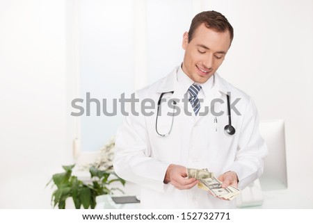Rich doctor. Cheerful young male doctor counting money at his working place