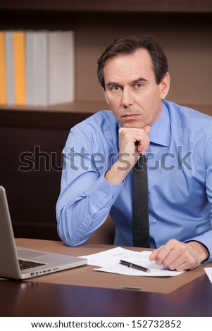 Confident businessman. Confident mature businessman sitting at his working place and holding his hand on chin