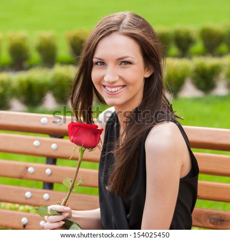 Girl with flower. Beautiful young woman holding red rose and smiling while sitting on the bench