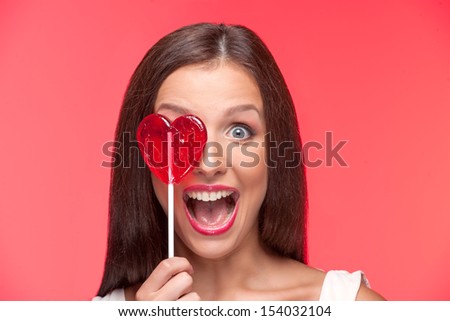 Girl with lollipop. Portrait of beautiful young woman holding heart shape lollipop in front of her eye while isolated on red