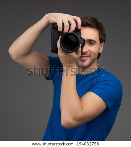 Man with camera. Cheerful young man holding camera in his hands and focusing at you while isolated on grey