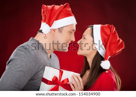 Together at Christmas Eve. Cheerful young couple in Santa hat holding a gift box and smiling to each other while standing isolated on red