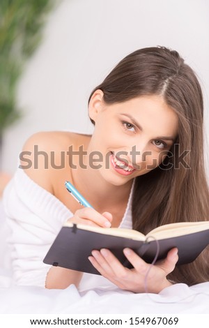 Making notes. Cheerful young woman writing something in her note pad and smiling while lying on the sofa