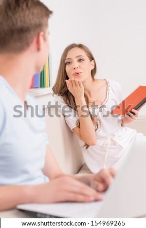 Loving couple at home. Cheerful young man working at the computer while his girlfriend holding book and blowing a kiss