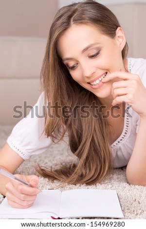 Girl with note pad. Cheerful young woman lying on the floor and writing something in her note pad