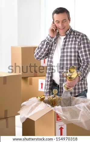 House moving. Cheerful middle-aged man packing the cardboard box and talking on the mobile phone