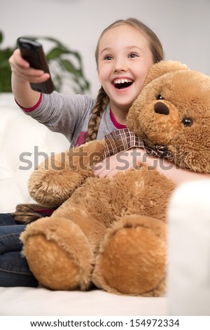 Cute girl at home. Cheerful little girl watching TV and holding toy bear