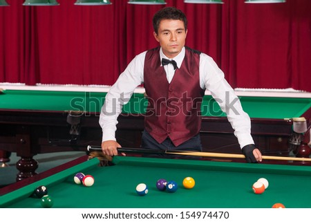 Pool player. Thoughtful young pool player holding cue and looking at billiard table