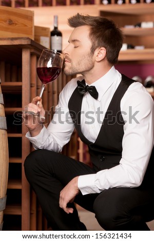 Confident sommelier. Thoughtful young sommelier smelling wine from the glass while sitting near wine barrel
