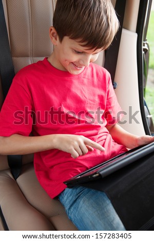Boy is sitting in a car and playing with tablet. Looking interested and smiling