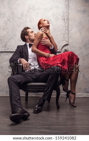 Elegant Handsome Man Kissing His Girlfriend In A Shoulder. Girl Sitting On His Knees