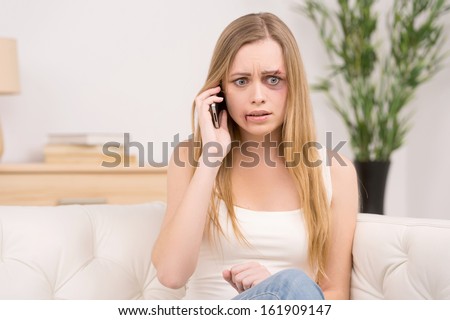 Scared woman with bruise on face calling to get help. Sitting on sofa in room