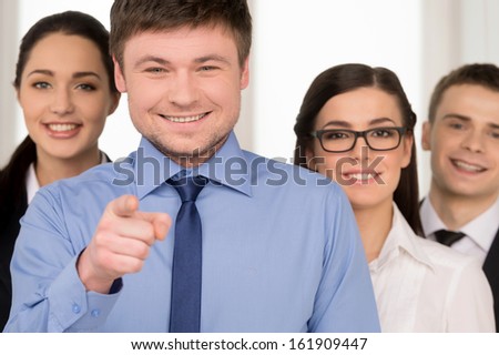Smiling successful business man pointing with finger on camera. Group of people standing on background
