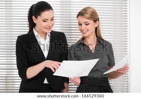 Two beautiful business woman sharing ideas with each other. Standing together and checking documents in modern office
