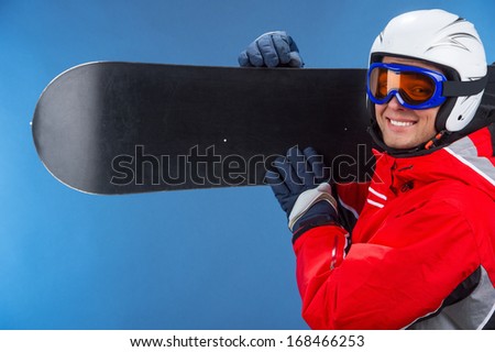 Close up of active smiling snowboarder with snowboard over shoulder. Looking at camera standing isolated over blue background