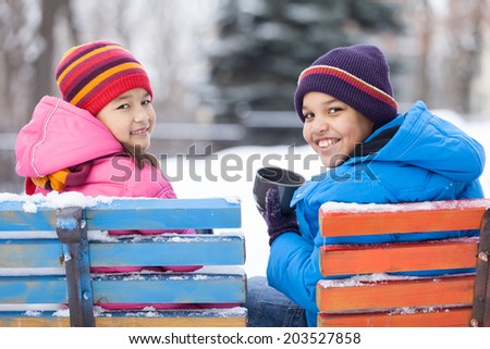 boy and girl sitting in park on bench. children smiling and drinking hot tea