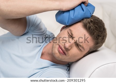 closeup of man holding icepack on his head. young man experiencing pain on white sofa