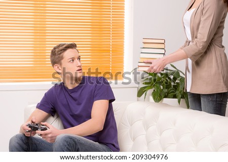 man playing computer game sitting on sofa. woman standing and holding stack of books
