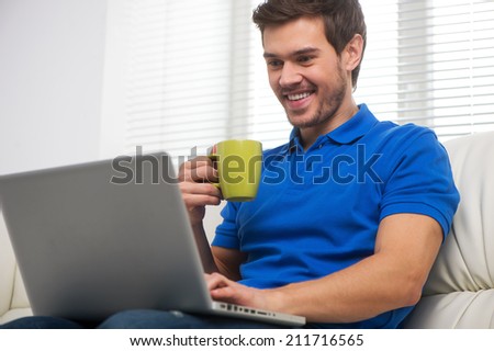 Young man happy on sofa with laptop. handsome male in t-shirt sitting on sofa and drinking