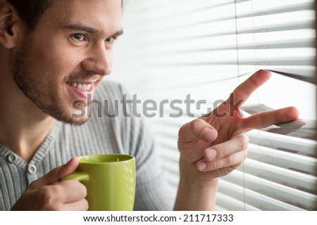 young man looking through blinds. Man looks through jalousie and holding tea cup