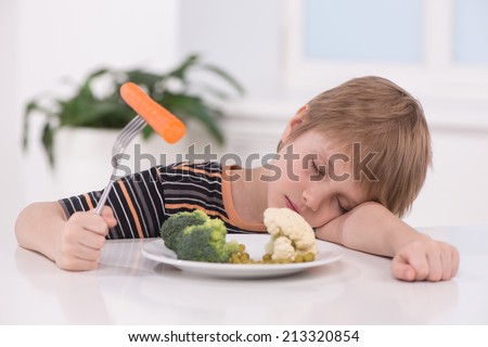 little blond boy eating at kitchen. child holding fork with carrot and sleeping