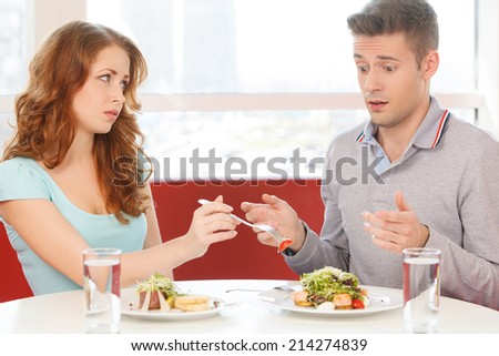 woman eating strawberry from man's plate. man sitting surprised and lifting hands in dismay