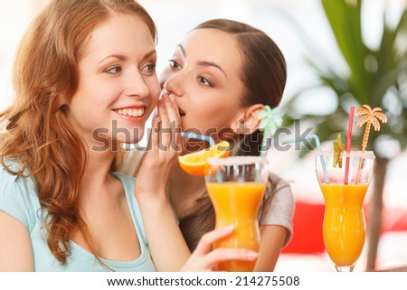 one woman whispering something to friend. two girls sitting in cafe and drinking juice