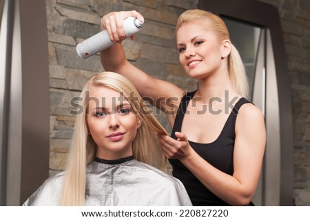 blond hairdresser applying spray on client's hair and looking into camera. Female hairdresser works on woman hair in salon