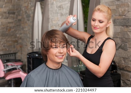 blond female hairstylist using hair spray. young attractive man sitting in hair salon and smiling