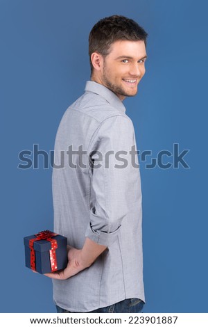 side view of smiling guy holding gift in blue box. waist up of happy male carrying blue box on blue background