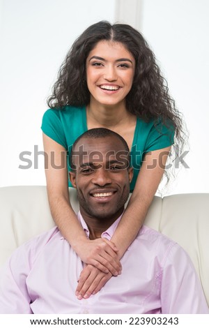Indian girl standing over man sitting on sofa and hugging. black african man sitting on sofa and smiling
